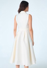 Robe A-line sans manches en tissu crème avec une taille ceinturée et une jupe plissée, présentant une silhouette ajustée et une texture lisse.