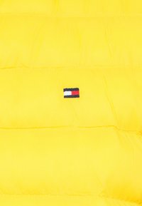 Gelbe Stepp-Wattierte Jacke mit gestepptem Design, versehen mit einem kleinen gestickten Logo in Blau, Weiß und Rot auf der linken Seite. Weiche, glatte Textur.