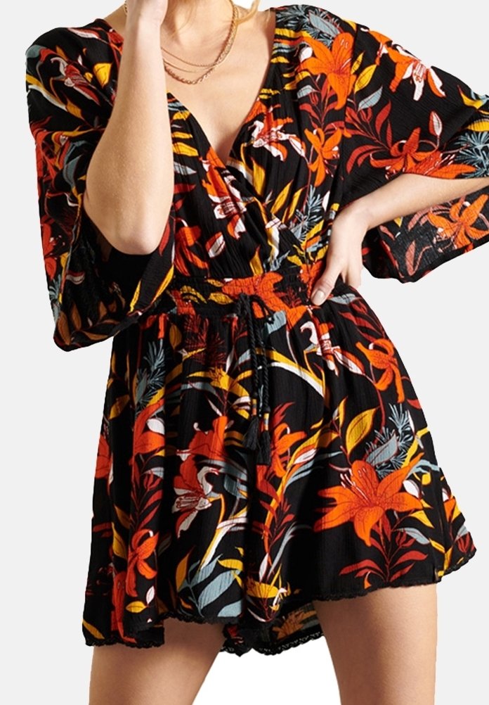 Femme portant une courte robe noire à motifs floraux orange, rouges et jaunes, manches bouffantes et taille cintrée avec un cordon.