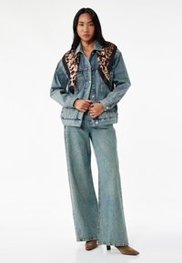 Chaqueta de denim con cuello de estampado de leopardo, lavado azul claro, corte oversize, combinada con pantalones de denim de pierna ancha. Tacones marrones de punta completan el look.