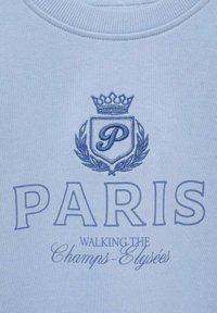 Ljusblå sweatshirt med broderad text: "PARIS" i fet stil, med "WALKING THE Champs-Élysées" nedanför, samt en krona och emblemdesign.