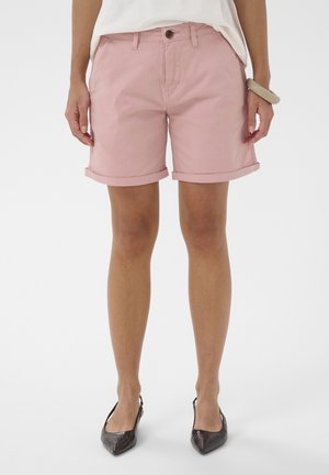 Vrouw die pastelroze shorts met opgerolde zoom draagt, een wit topje, bruine slingback schoenen met puntige neus en een beige armband om haar pols.