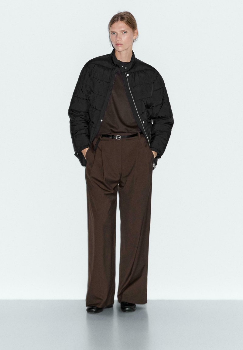 Woman wearing a black quilted jacket over a dark brown top tucked into high-waisted wide-leg brown pants with black shoes.