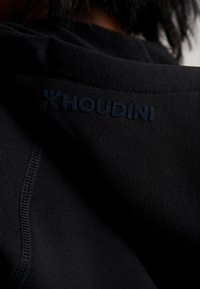 Musta kangashuppari, jossa on sileä pinta, ja joka näyttää tikkauksella ommellun logon "HOUDINI" tummansinisellä kaulan alueella. Yksinkertainen muotoilu, jossa on vähän yksityiskohtia.