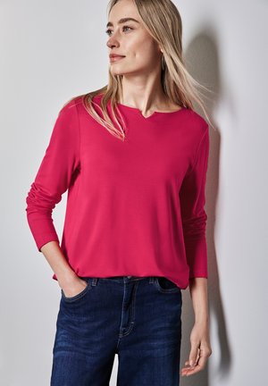Růžový top s dlouhými rukávy a kulatým výstřihem, hladká textura, volný střih, kombinovaný s tmavě modrými džíny. Jednoduchý a moderní design.