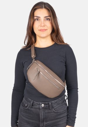 Frau trägt eine taupefarbene Leder-Umhängetasche über einem schwarzen Langarmshirt und dunkelgrauen Jeans vor einem schlichten Hintergrund.