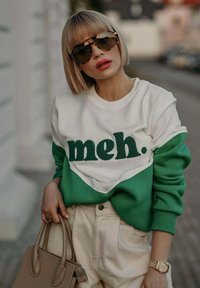 Mikina s blokováním barev v bílé a zelené barvě, s textem "meh." v strukturované zelené. V kombinaci s béžovými kalhotami s vysokým pasem a kabelkou.