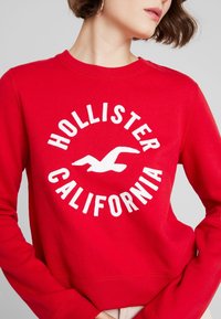 Sudadera roja con un gráfico circular blanco que incluye el texto "HOLLISTER" y "CALIFORNIA", fabricada de una mezcla de algodón, con puños y dobladillo acanalados.