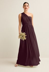 Femme portant une robe longue asymétrique violet foncé tenant un petit bouquet de fleurs séchées, debout sur un fond clair.