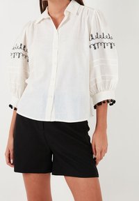 Blouse blanche à boutons avec des accents brodés noirs et des manches bouffantes, dotée d'une finition en pompons noirs au niveau des poignets. Associée à un short noir.