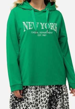 Person iført grøn hættetrøje med "New York Casual Department Est. 1987" print og sort-hvid leopardprint nederdel.