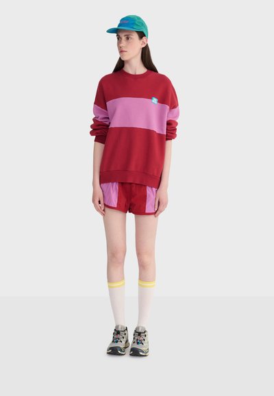 Jeune personne portant un sweat-shirt à blocs de couleurs rouge et rose, un short, des chaussettes blanches montantes avec des rayures jaunes, des baskets grises et une casquette turquoise.