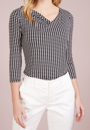 Top a maniche lunghe a scacchi bianco e nero con scollo a V e dettaglio arricciato al collo, abbinato a pantaloni bianchi.