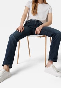 Jeans de mezclilla azul oscuro con diseño de pierna recta, combinados con una camiseta blanca sencilla y zapatillas blancas, sentados en una silla de madera.