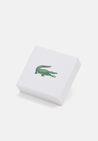 Kvadratinė balta dėžutė su tekstūruotu paviršiumi, ant kurios matyti žalias krokodilo logotipas. Paprastas dizainas, švarios kraštinės, minimalistinė išvaizda.