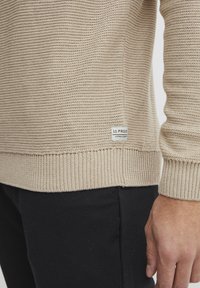 Beige kötött pulóver bordázott mandzsettával és szegéllyel, texturált mintával és egy kis márkajelzéssel az alsó oldalán.