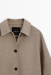 Beige wolmix jas met een puntige kraag, zichtbare knopen en een gestructureerde afwerking. Etiket is zichtbaar aan de binnenkant met de merknaam.
