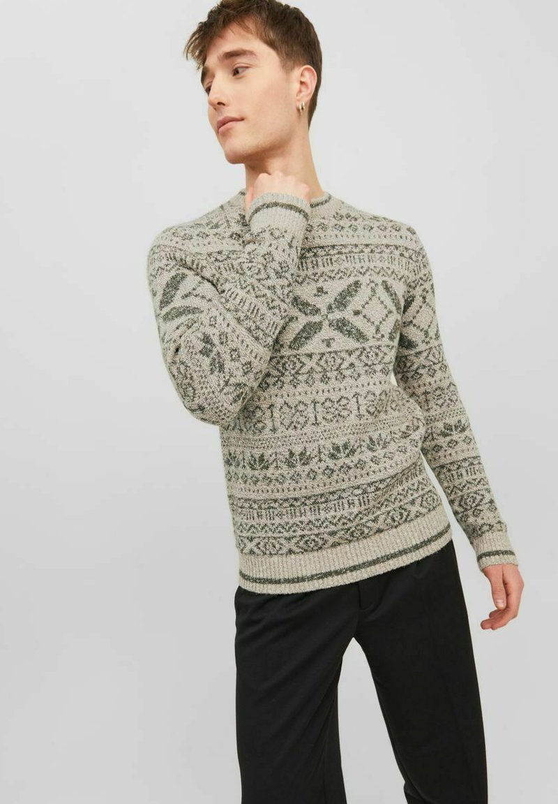 Strickpullover in Beige mit grünen geometrischen und floralen Mustern, Rundhalsausschnitt und gerippten Bündchen. Kombiniert mit schwarzen Hosen.