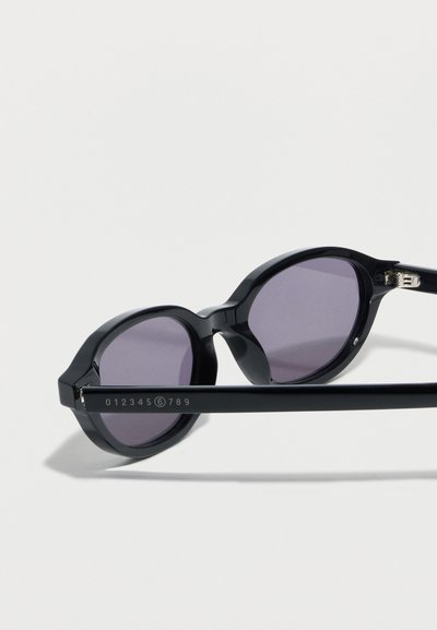 Μαύρα στρογγυλά γυαλιά ηλίου με μοβ φακούς που είναι τοποθετημένα πλαγίως, δείχνοντας το μπράτσο του σκελετού με αριθμούς, συμπεριλαμβανομένου ενός κύκλου με το 6.