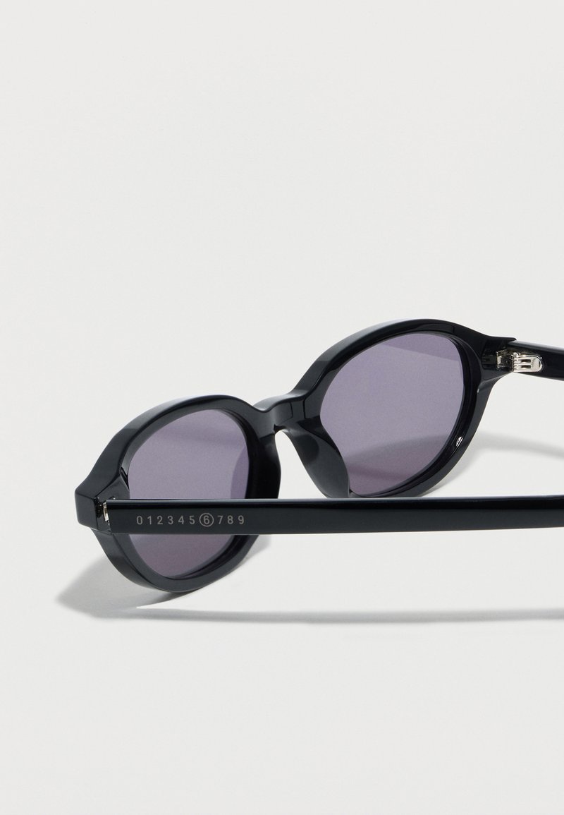 Gafas de sol redondas negras con lentes moradas, acostadas de lado, mostrando la varilla marcada con números, incluyendo un 6 dentro de un círculo.