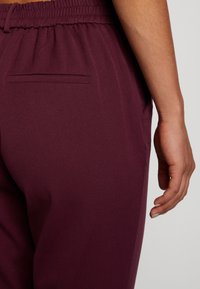 Burgundowe joggery z miękkiego, fakturowanego materiału, z elastycznym pasem i jedną tylną kieszenią, częściowo widoczna ręka spoczywająca obok.