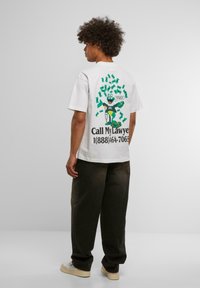 Hvid bomulds t-shirt med et farverigt tegneseriefigur, der har dollarsedler og tekst på ryggen. Løs pasform parret med mørke bukser.