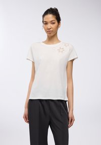 T-shirt bianco in cotone a maniche corte, con ricami intricati a forma di stelle sulla spalla sinistra. Abbinato a pantaloni neri.