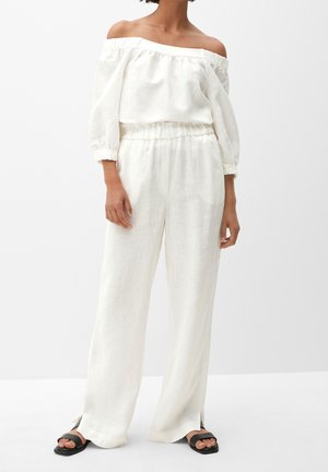 Frau trägt weiße Bluse mit Carmenausschnitt und Puffärmeln sowie passende weite Hose, kombiniert mit schwarzen Sandalen zum Hineinschlüpfen.