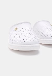 Sabots blancs avec de petites perforations noires, texture lisse, forme arrondie et un accent doré sur le côté. Semelle souple et légère.