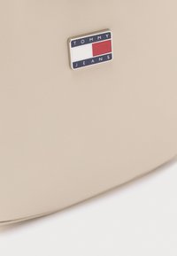 Bézs színű kézitáska puha anyagból, sima felülettel, kék, fehér és piros színű márkajelzéssel ellátott táblával.