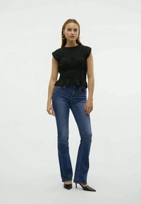 Schwarzes Peplum-Oberteil mit kurzen Ärmeln, kombiniert mit eng anliegenden blauen Jeans. Das Model trägt schwarze Stilettos. Neutraler Hintergrund.
