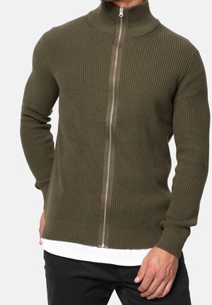 Homme portant un pull zippé côtelé vert olive avec un col montant, un t-shirt blanc et un pantalon noir sur un fond uni.