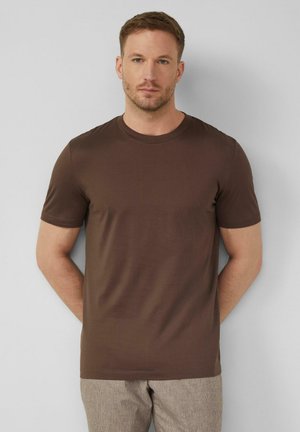 Mężczyzna z krótkimi włosami i jasnym zarostem nosi gładki brązowy krótki rękaw t-shirt i beżowe spodnie na tle szarej ściany, stoi z rękami za plecami.