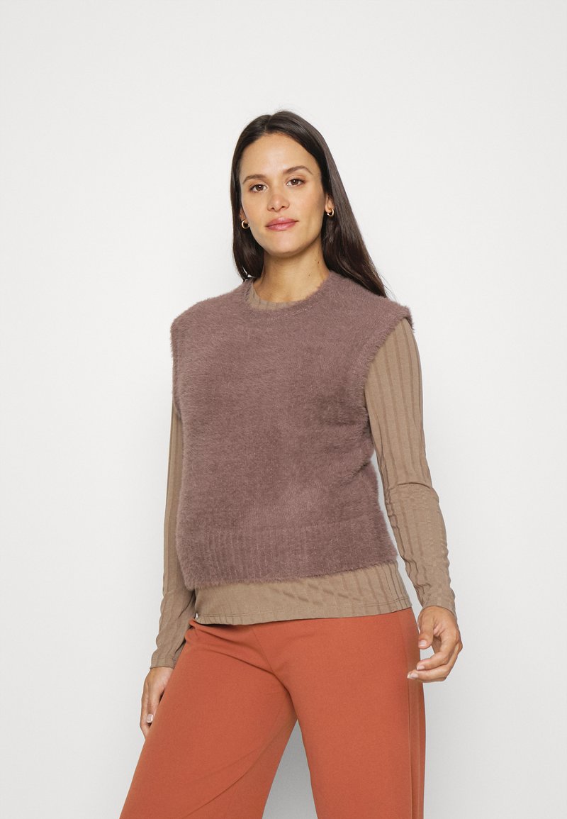 Colete de malha fuzzy em malva, com decote redondo e design cropped, sobre uma blusa de manga longa bege com padrão canelado e calças laranja.