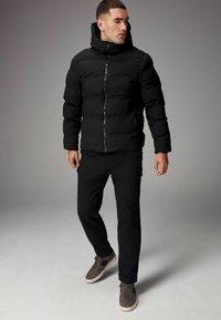 Sort pufferjakke med høj krave, lynlås-lukning og sidelommer; pared med sorte bukser og grå sneakers. Glat tekstur overalt.