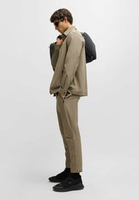 Vaaleanruskea kevyt takki ja siihen sopivat housut vetoketjuilla, yhdistettynä mustiin lenkkareihin. Mallilla on musta reppu, ja hän seisoo yksinkertaista taustaa vasten.