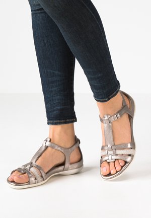 Metallic bronze sandals with an open design, featuring crisscross straps and a cushioned sole, paired with dark skinny jeans.