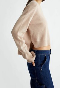 Nainen, jolla on yllään beige lyhyt neuletakki, jossa on ribbimaalit, ja siniset korkeavyötäröiset farkut, joissa on suuret sivutaskut ja napitus.