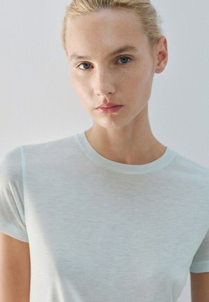 Jonge vrouw met lichtblond haar in een staart gebonden, die een eenvoudig wit T-shirt draagt en rustig in de camera kijkt tegen een lichte achtergrond.