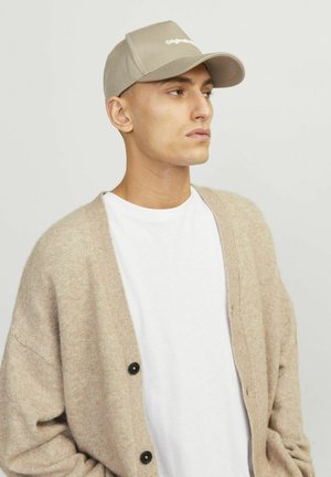 Casquette beige avec une visière incurvée, logo brodé, et cardigan beige en tissu texturé avec des boutons noirs sur une chemise blanche.