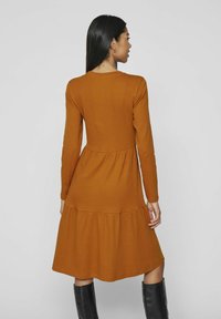 Långärmad, rostfärgad klänning med en figurnära bodice och utsvängd kjol. Ribbad textur, lagerpappa, och rund halsringning.