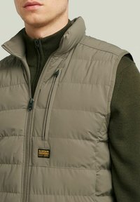 Gilet imbottito color khaki con collo alto, dotato di tasca con zip, texture trapuntata e patch logo G-Star Originals sul lato.