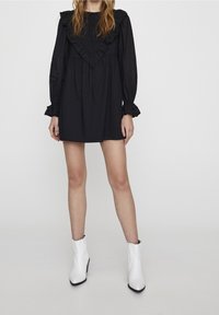 Robe noire avec un col à volants, manches longues et taille froncée. Associée à des bottines blanches à bout pointu et fermeture éclair latérale.