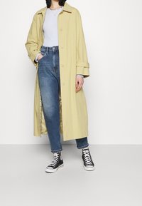 Žlutý trench coat s volným střihem, zapínáním na knoflíky a širokými manžetami, nošený přes světle modrý žebrovaný top, v kombinaci s vysokým pasem modrými džíny a černými teniskami.