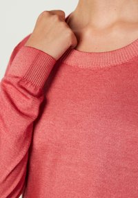 Korallowy sweter dziany z okrągłym dekoltem i prążkowanymi mankietami. Materiał wydaje się miękki, o subtelnej fakturze i lekkiej konstrukcji.