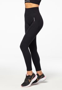 Black high-waisted leggings with ribbed side panels, stretchy fabric, and a logo detail on the hip. Paired with black athletic shoes.