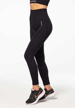 Black high-waisted leggings with ribbed side panels, stretchy fabric, and a logo detail on the hip. Paired with black athletic shoes.