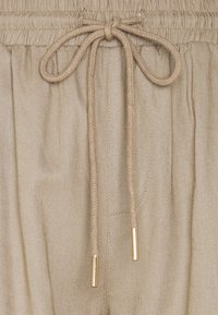 Beige drawstring tailleband met elastische details, voorzien van gevlochten koorden met goudkleurige metalen aglets, getoond tegen een textuurstof achtergrond.
