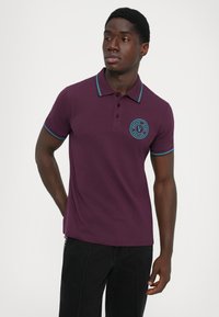 Νεαρός άνδρας που φοράει ένα μωβ πόλο μπλουζάκι με τιρκουάζ λεπτομέρειες και λογότυπο Versace Jeans Couture, στέκεται με το ένα χέρι πίσω από την πλάτη του.