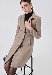 Cappotto doppio petto beige in misto lana con bottoni dorati, revers a punte e tasche laterali. Modella seduta su uno sgabello nero.